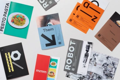 Flat lay of various graphic design publications, zines, and printed booklets with bold typography and imagery on a neutral surface.