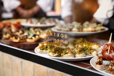 Plates of assorted pintxos and tapas displayed on a counter with chefs working in the blurred background.
