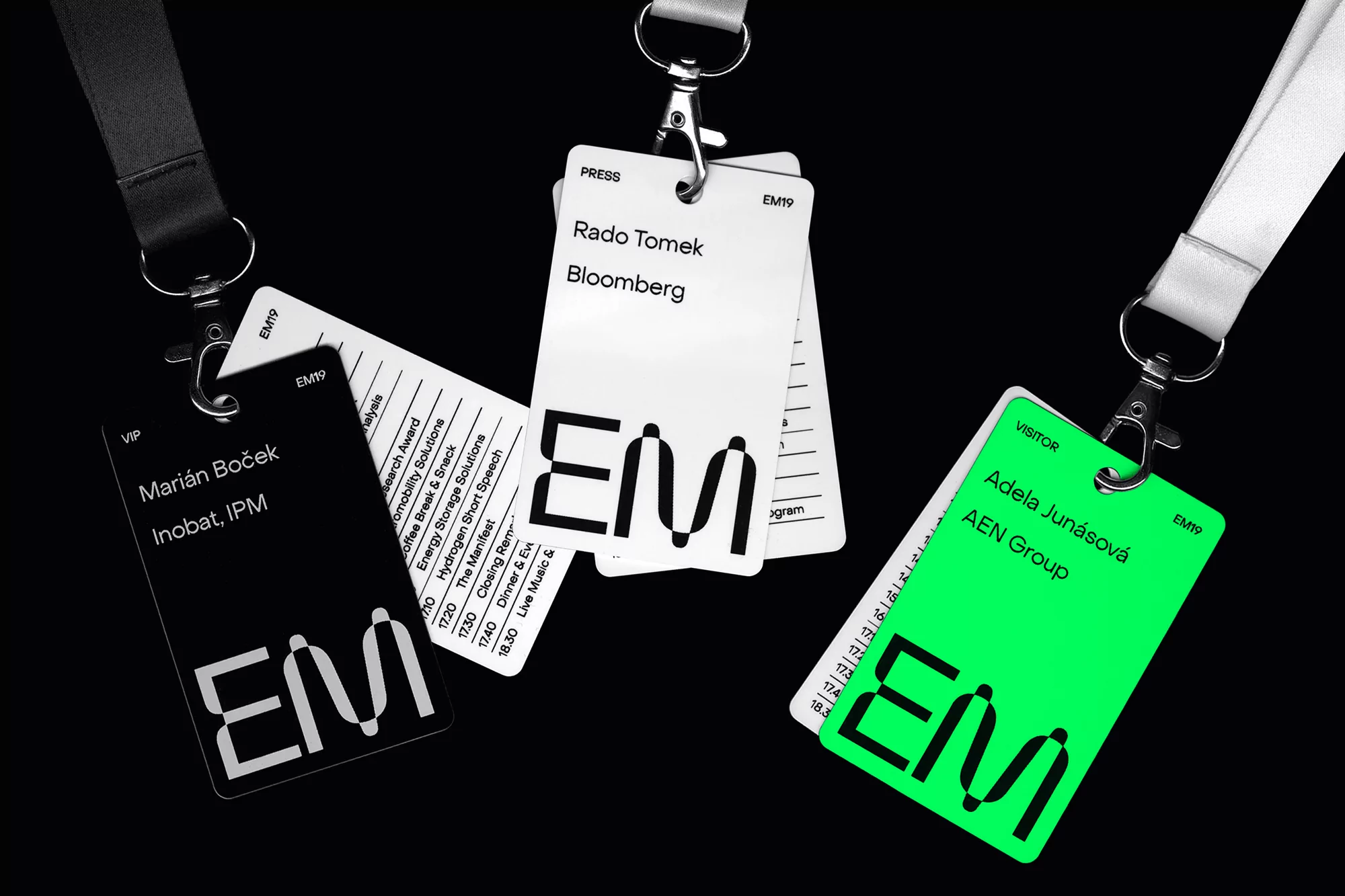 Three event badges on lanyards in black, white and green, with the logo 'EM' and event attendee names displayed on a black background.