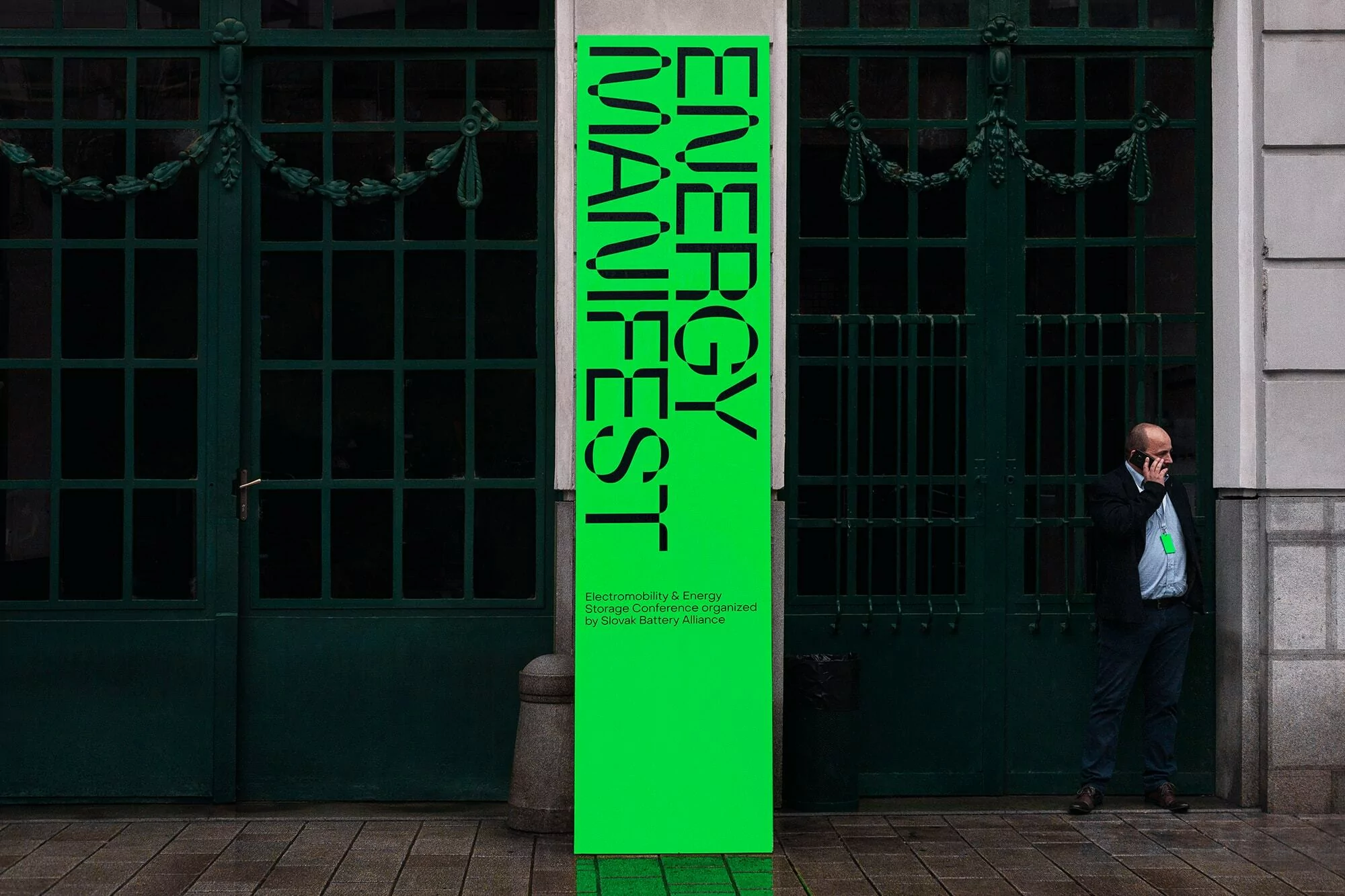 Tall banner with the words 'Energy Manifest' in black, standing on a sidewalk in front of large green doors, with an adult male on phone.