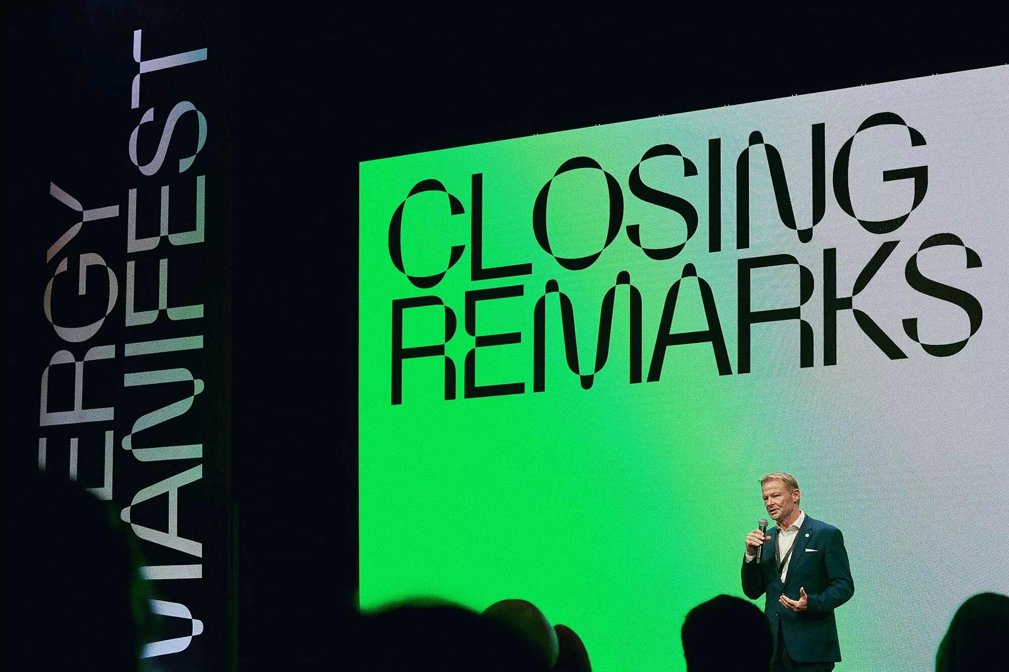 A man in a suit speaks on stage with a microphone in front of a large LED screen displaying closing remarks to an audience.
