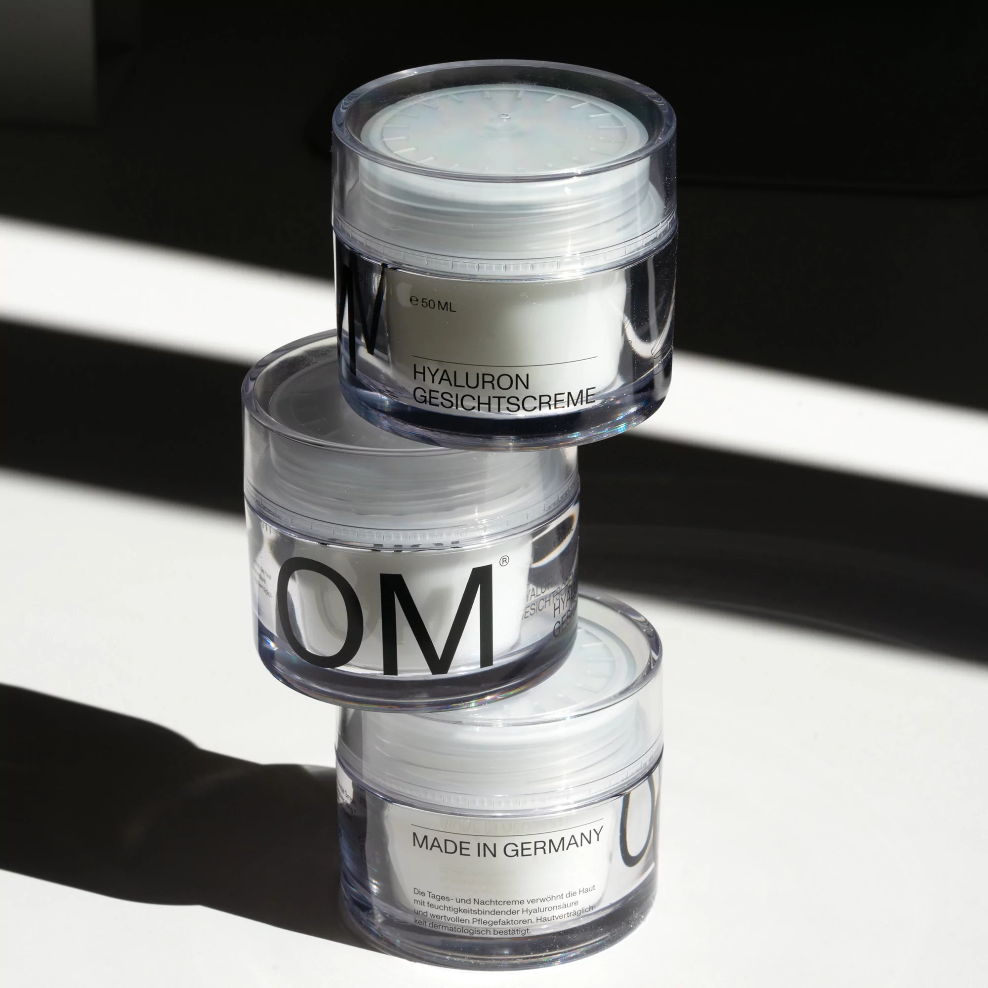Three clear jars of white face cream stacked on each other, casting shadows on a white surface, product shot for beauty advertisement.