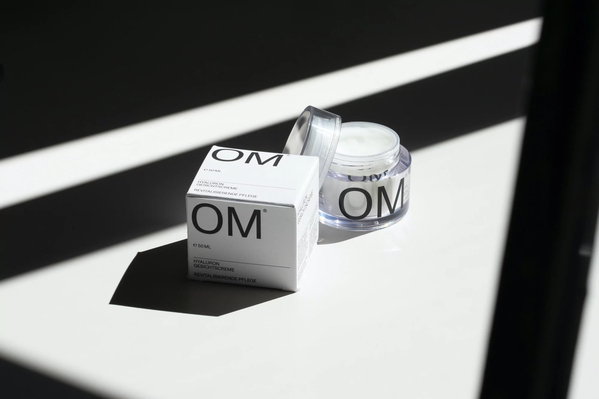 Cosmetic product presentation featuring a white box and open jar of face cream with modern, minimalist branding in a brightly lit setting.