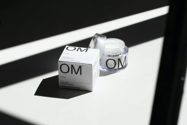 OM skincare product packaging and open cream jar with stark diagonal light and shadow patterns on a white surface.