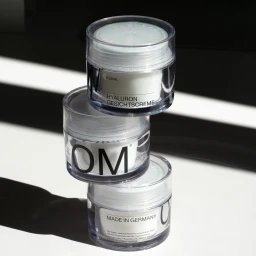 Three stacked clear skincare jars with white labeling against a white background under hard light.