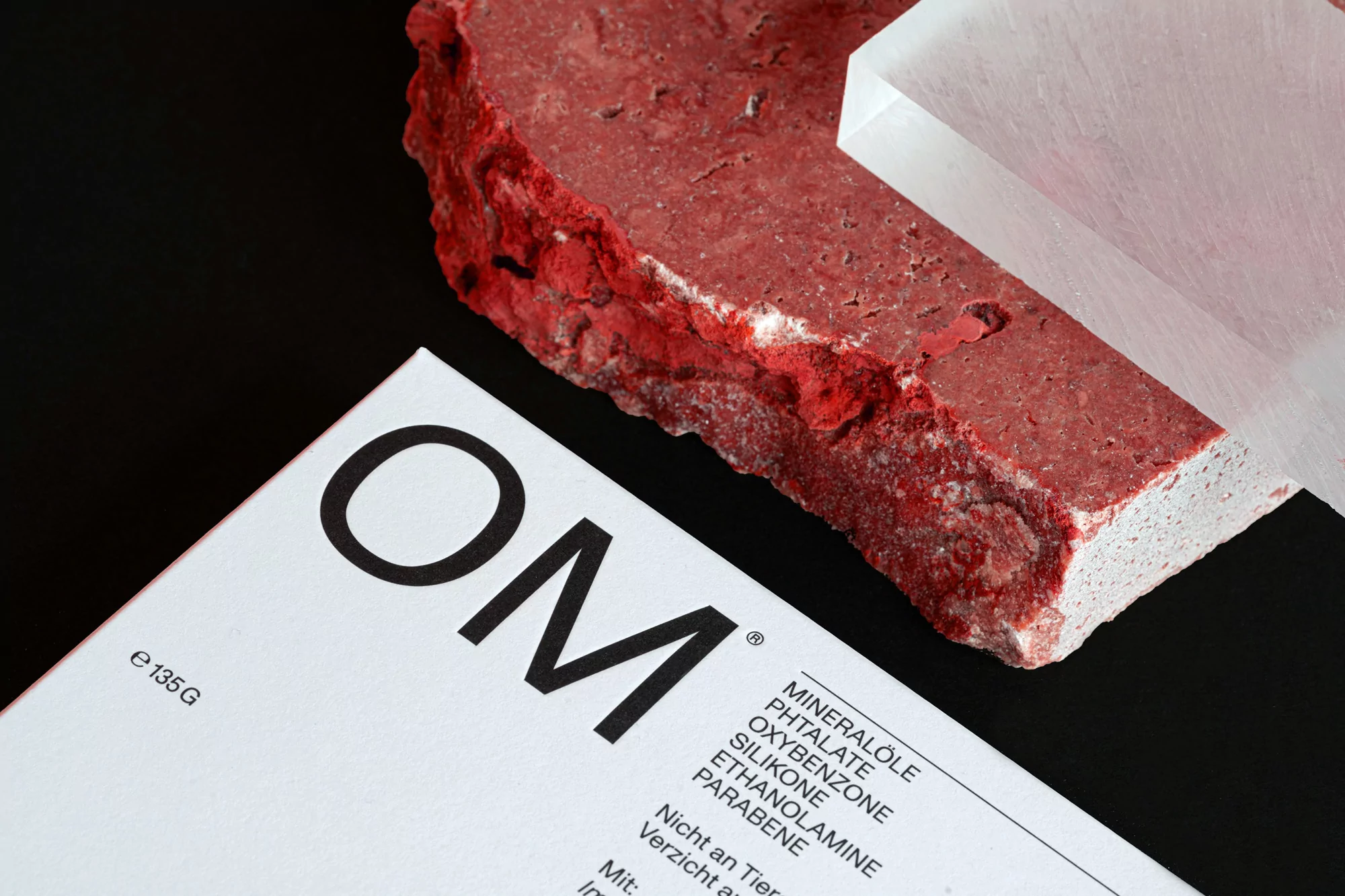 OM brand product packaging next to textured red stone and a transparent acrylic block on black background