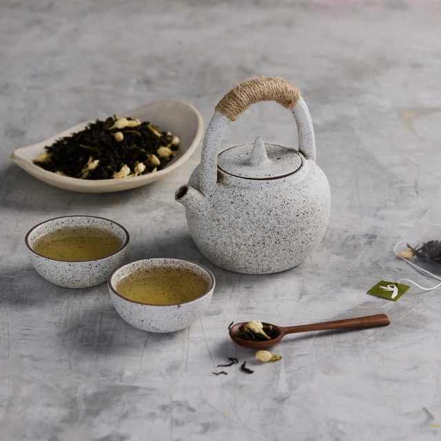 Speckled teapot with a woven handle next to two filled teacups, a small dish with tea leaves, and a wooden spoon on a gray surface.