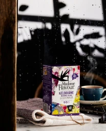 Madame Flavour tea box resting on folded fabric with a steaming mug behind a rain-streaked window