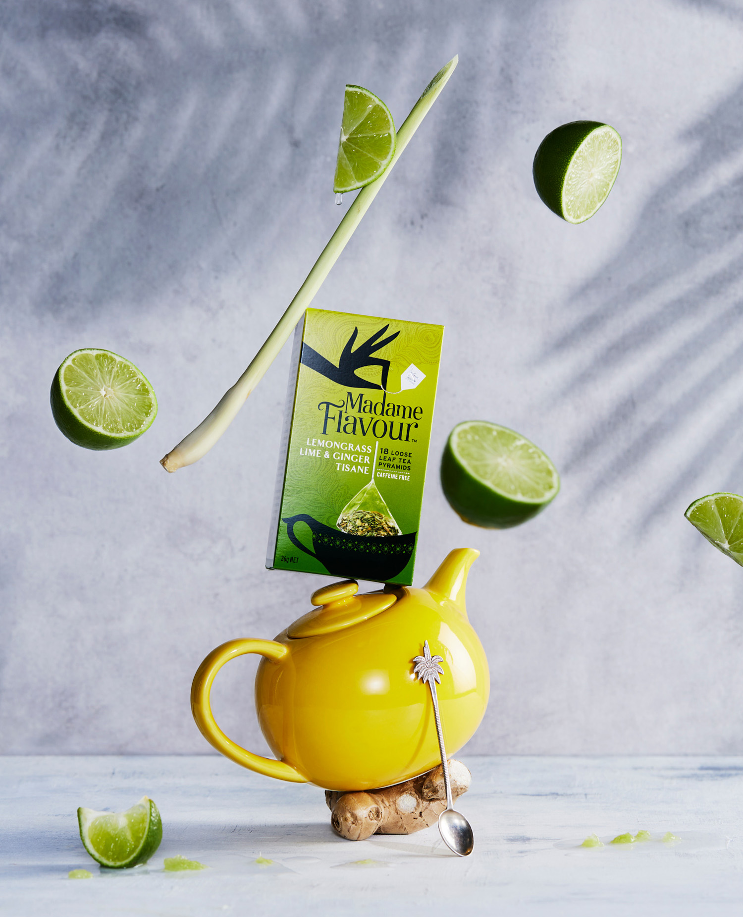 Still life showcases a teapot, limes, lemongrass, and tea packaging against a gray background with plant shadows, creating a fresh, vibrant food scene.