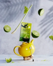 Madame Flavour tea box balanced on a yellow teapot with floating lime halves and lemongrass against a light textured background