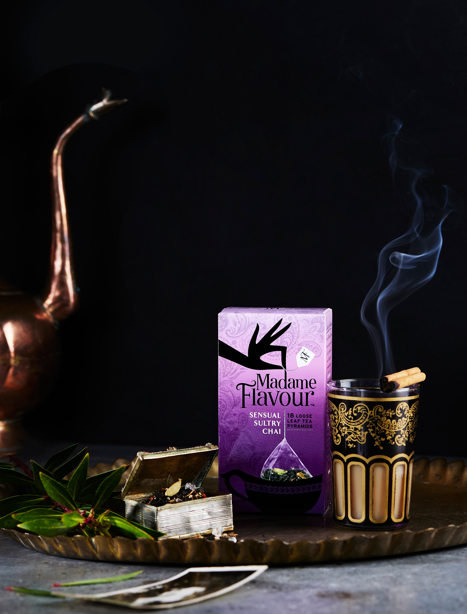 Still life featuring Madame Flavour Sensual Sultry Chai tea box, ornate cup with cinnamon sticks, silver box, and bay leaves on metal tray.