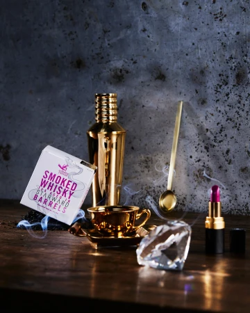 Still life shot on wooden table with golden bar tools, lipstick, and a box of cigars in a dark photo studio setting.
