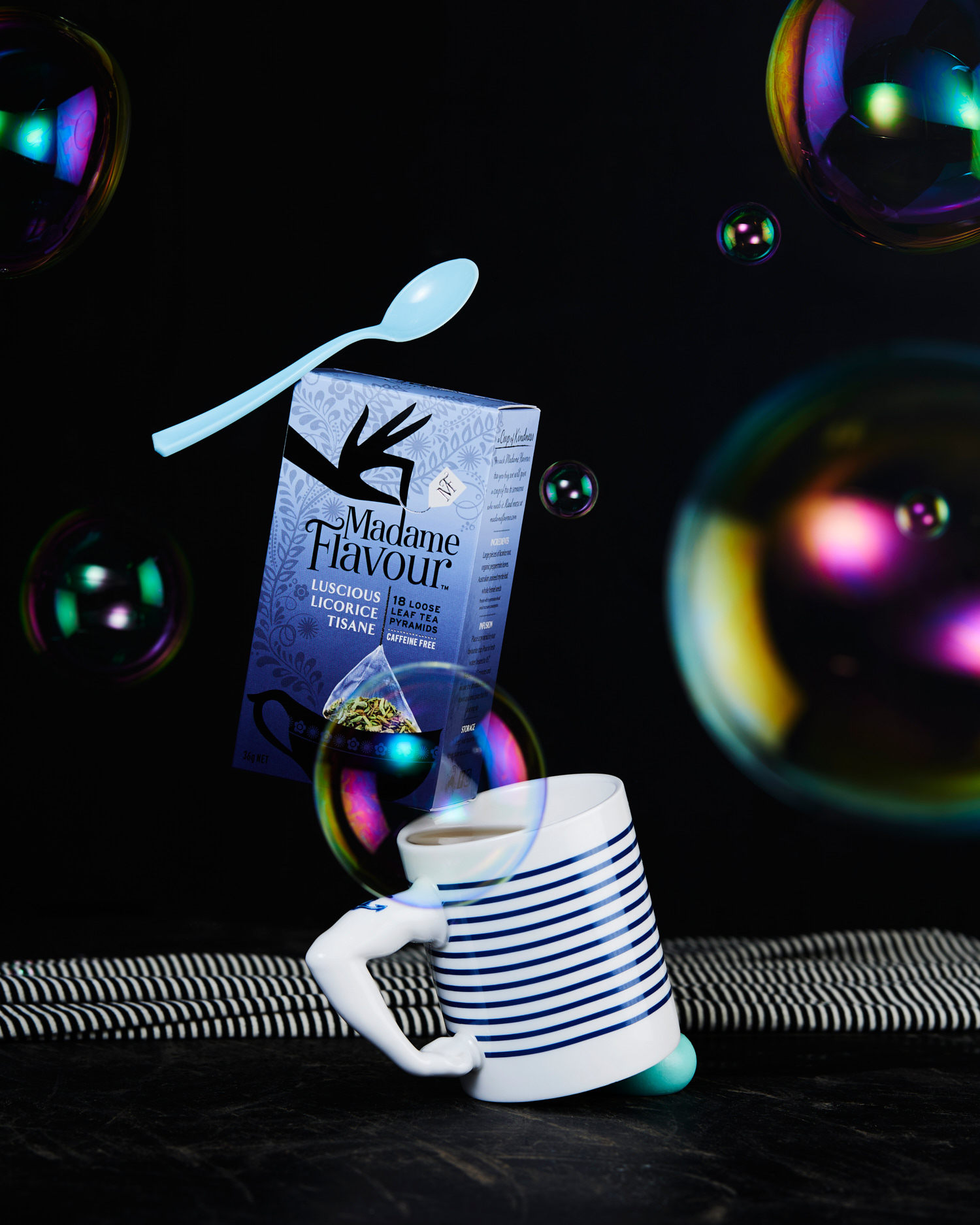 Still life photograph of a mug, tea box, and floating spoon, set against a dark background with several transparent soap bubbles.