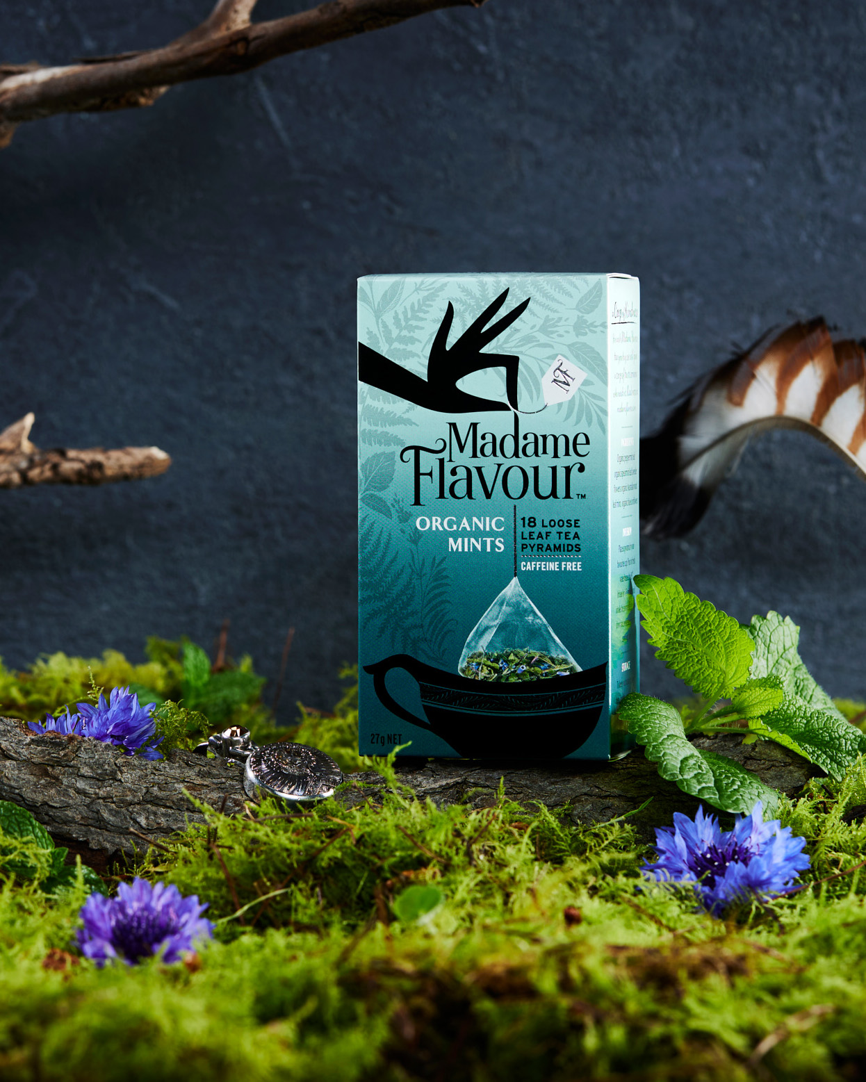 Madame Flavour Organic Mints tea box sits on moss with purple flowers and leaves against a dark blue background in a studio shot.