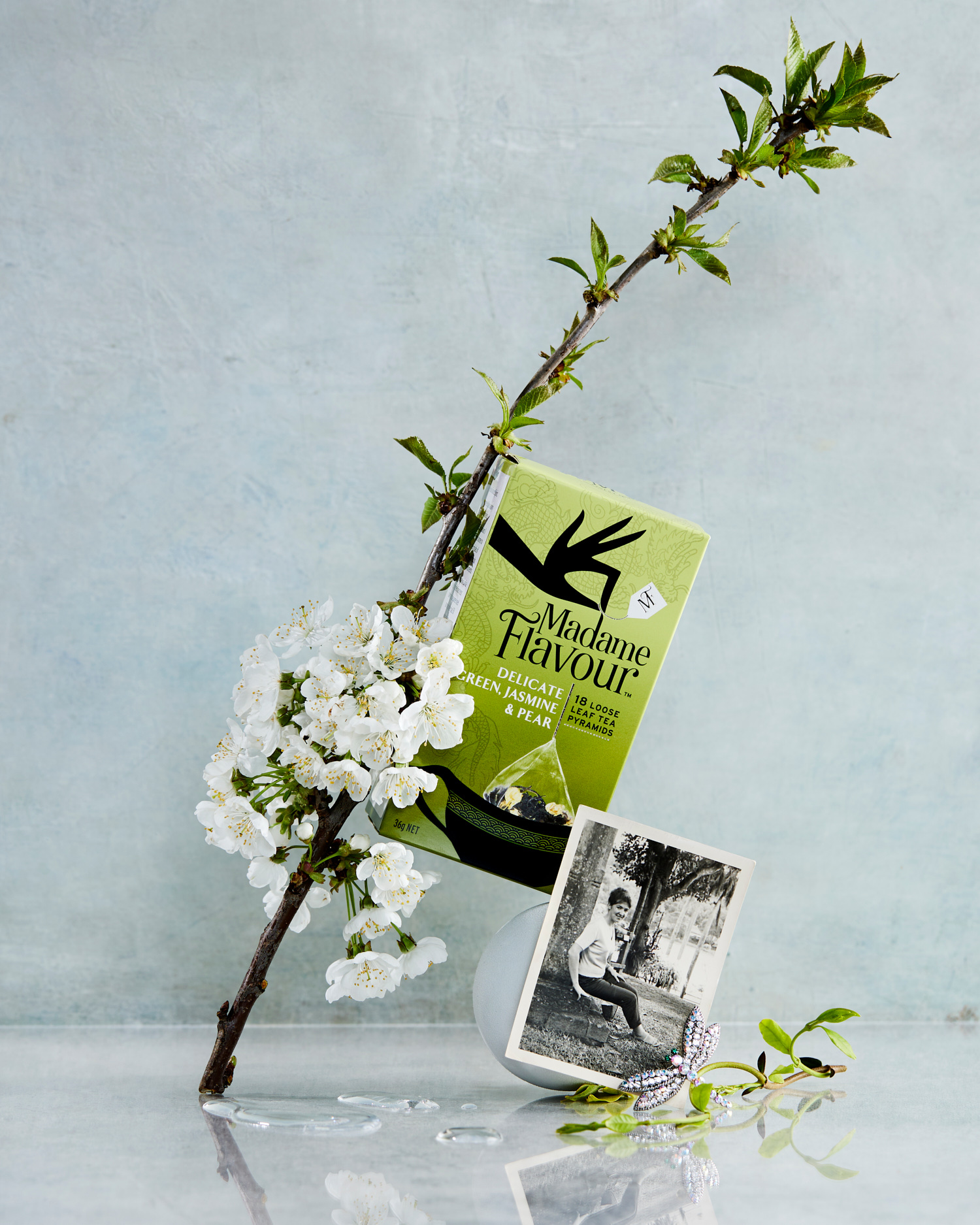 Still life with Madame Flavour tea box, white flowers on a branch, and a small framed black and white photo on a reflective surface.