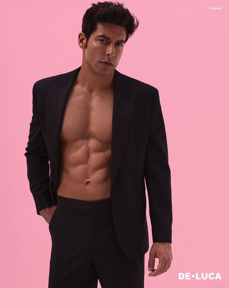 A muscular man poses in a black suit jacket and matching trousers against a light pink backdrop in a fashion shot. The man's torso is bare.