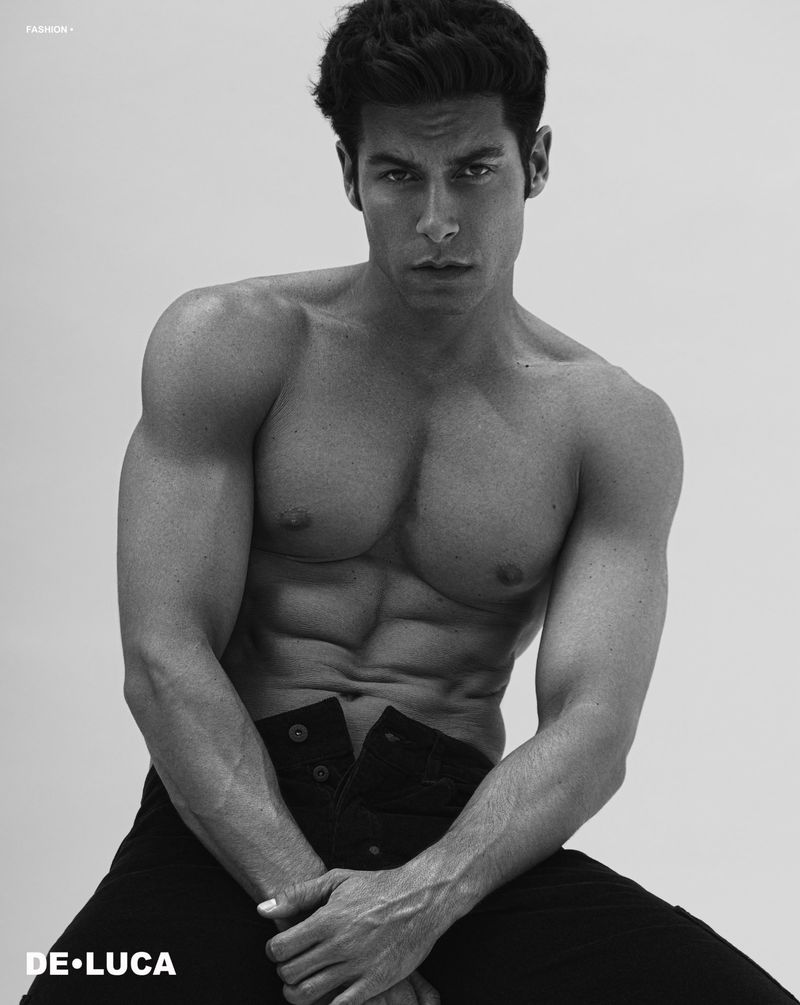 Black and white studio portrait of a shirtless adult male with dark hair sitting against a plain background, wearing dark denim pants.