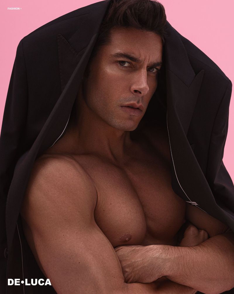 Nude male model poses with a black jacket draped over his head against a pink background in a fashion photoshoot.