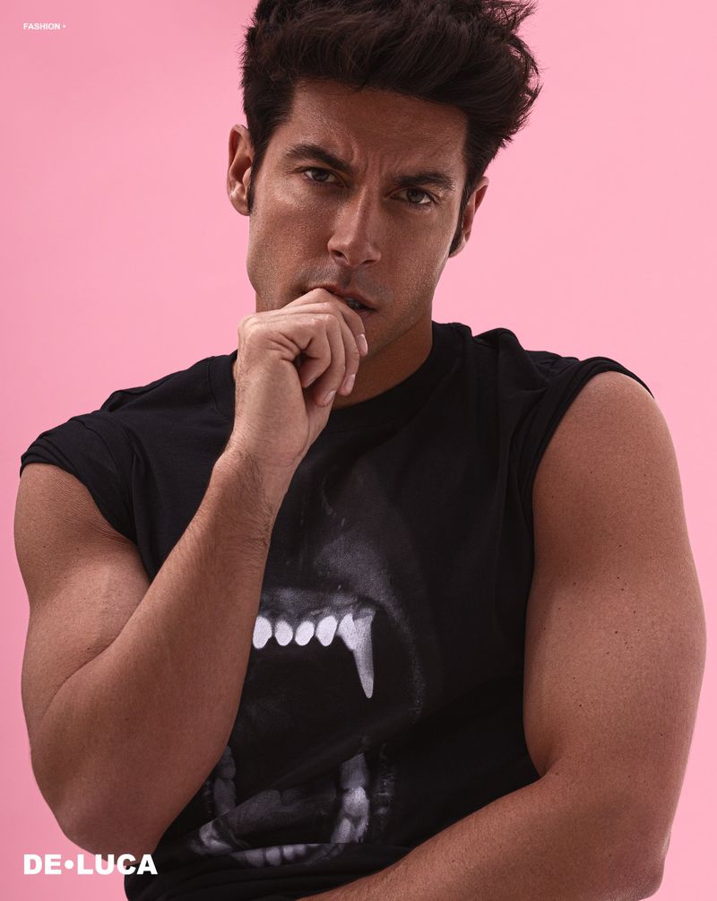 Portrait of a muscular adult man with dark hair and a black t-shirt, posing in front of a pink backdrop. He has his hand to his chin.