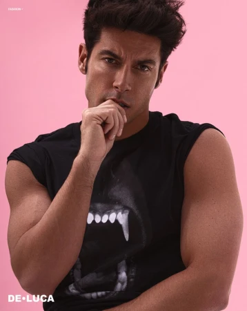 Portrait of a muscular adult man with dark hair and a black t-shirt, posing in front of a pink backdrop. He has his hand to his chin.