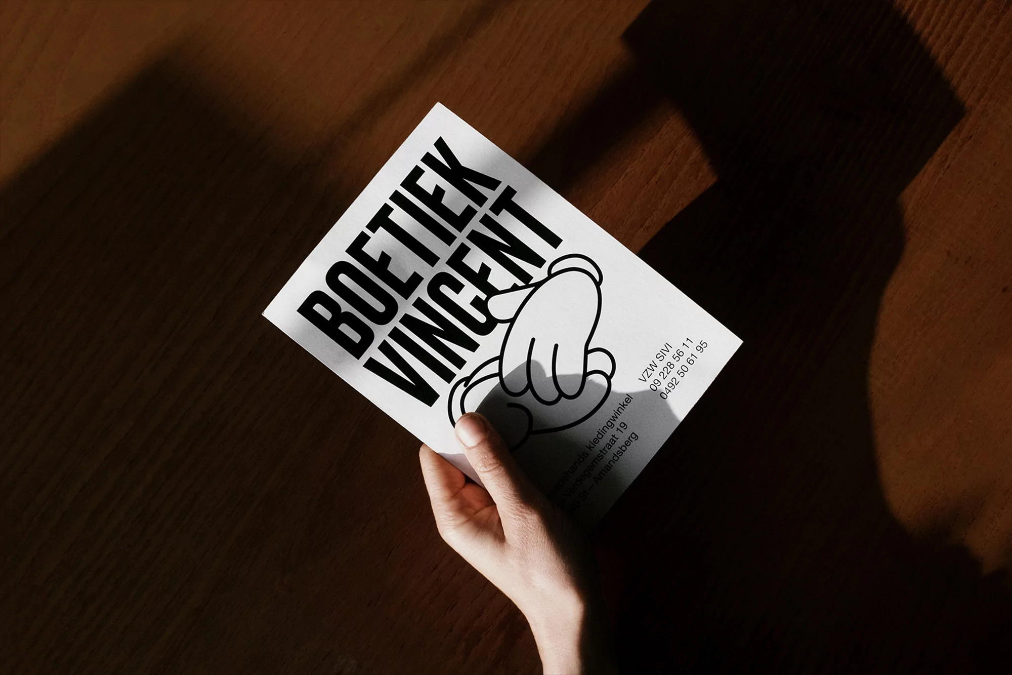 Person holding a white printed card on a wooden surface, featuring black text, cartoon illustration, and contact information.