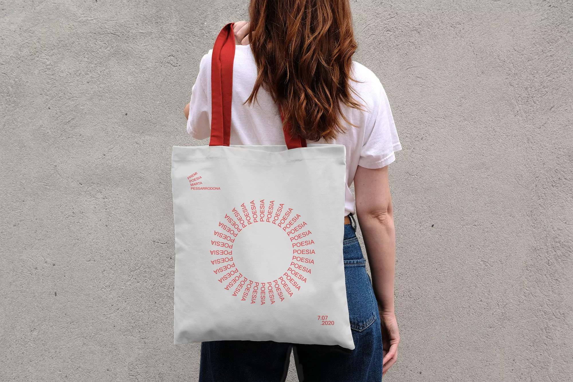 Back view of a woman holding a white tote bag with red handles and red text design, wearing a white shirt and jeans against a wall.