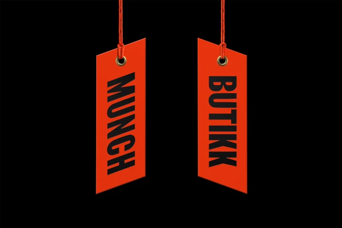 Two vibrant orange tags hang against a stark black background, each labeled 'MUNCH' and 'BUTIKK' in bold, sans-serif black text.