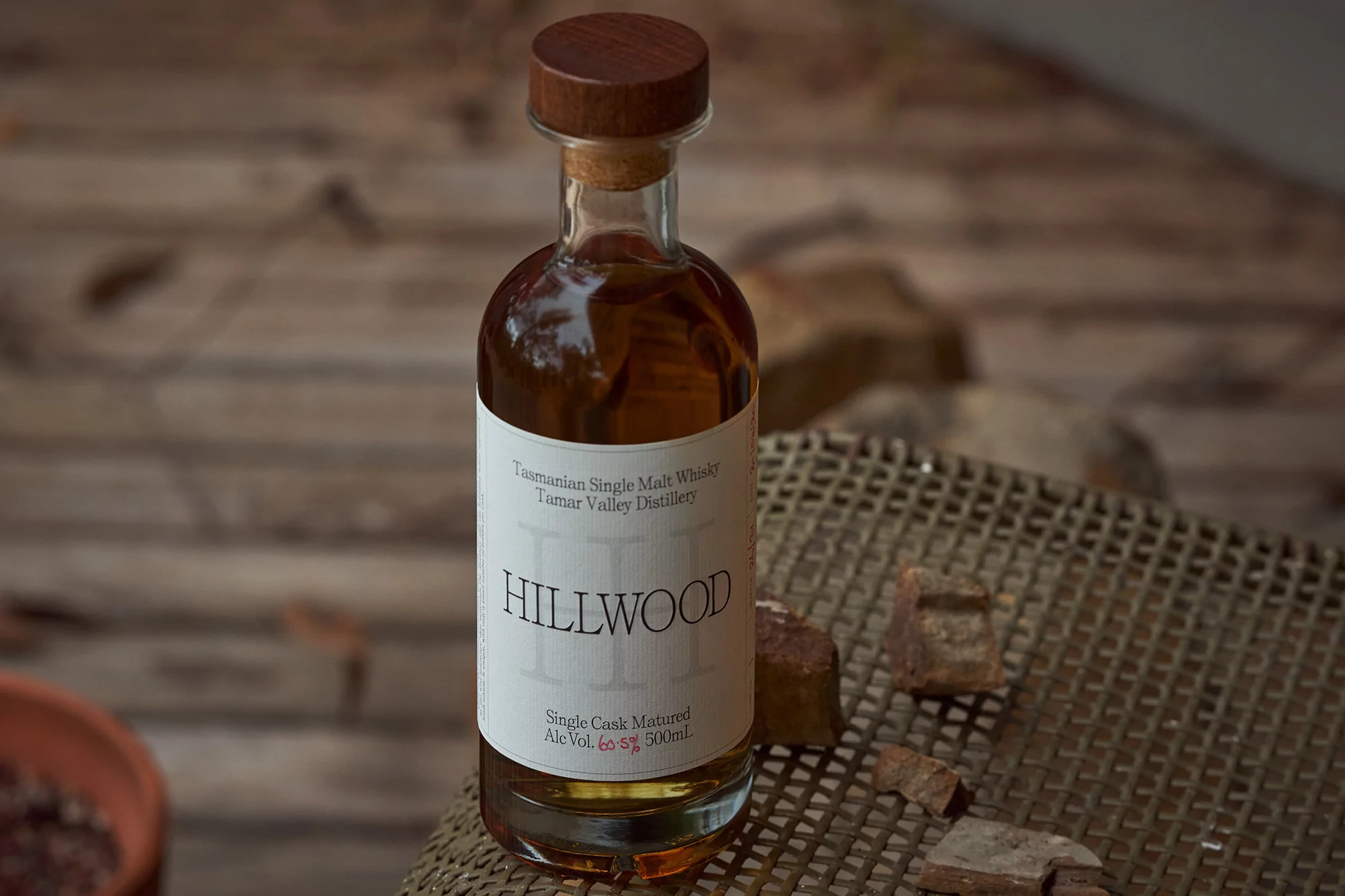 Whiskey bottle of Hillwood Tasmanian Single Malt Whisky with cork and wooden cap on rattan surface and rock fragments, rustic charm.