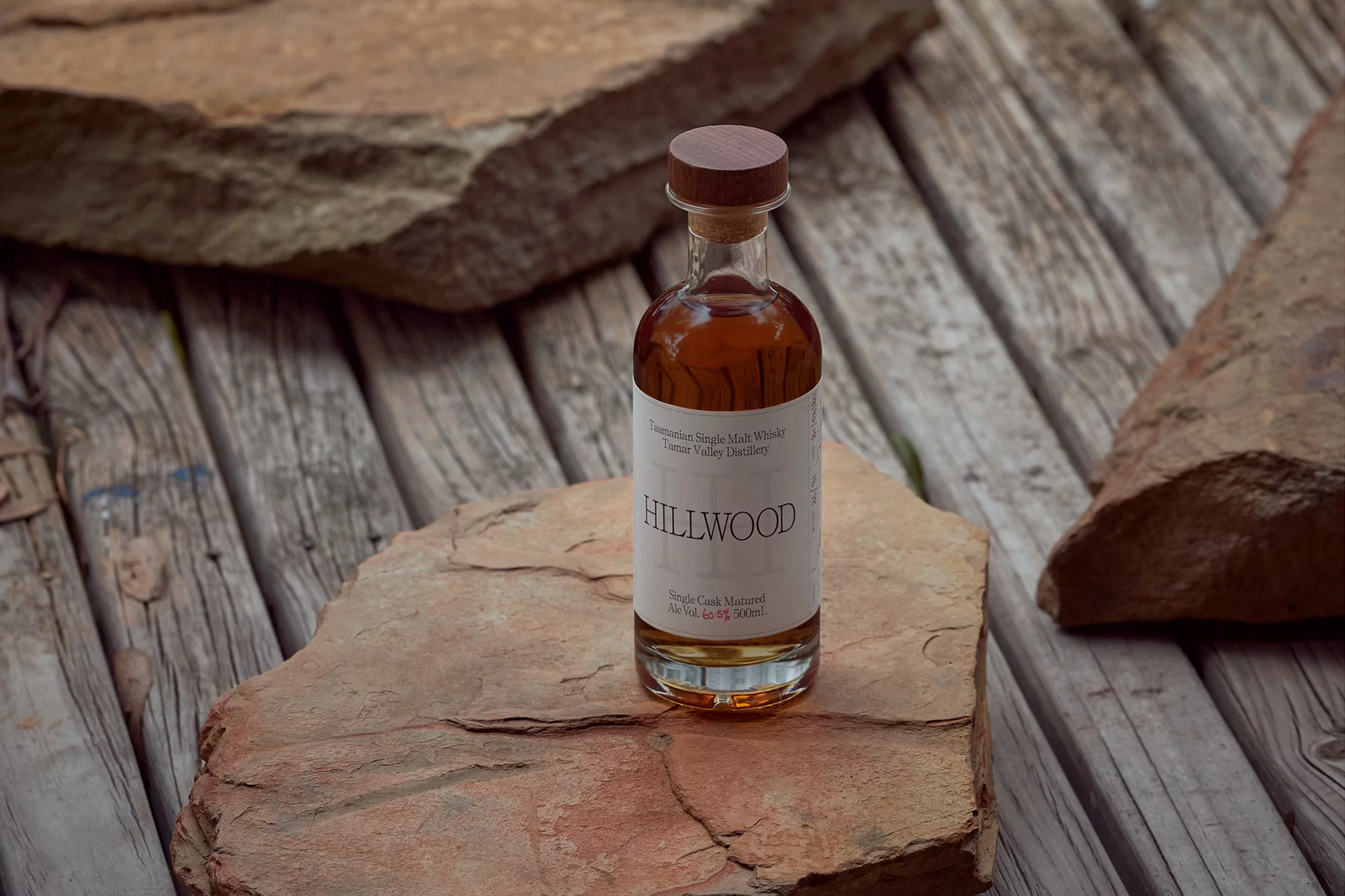 Whiskey bottle centered on stone surface with wooden planks, presenting natural textures and amber hues in a product shot.