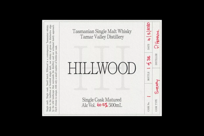 Close-up of a white paper product label for Hillwood Tasmanian Single Malt Whisky with black typography and handwritten red ink notations.
