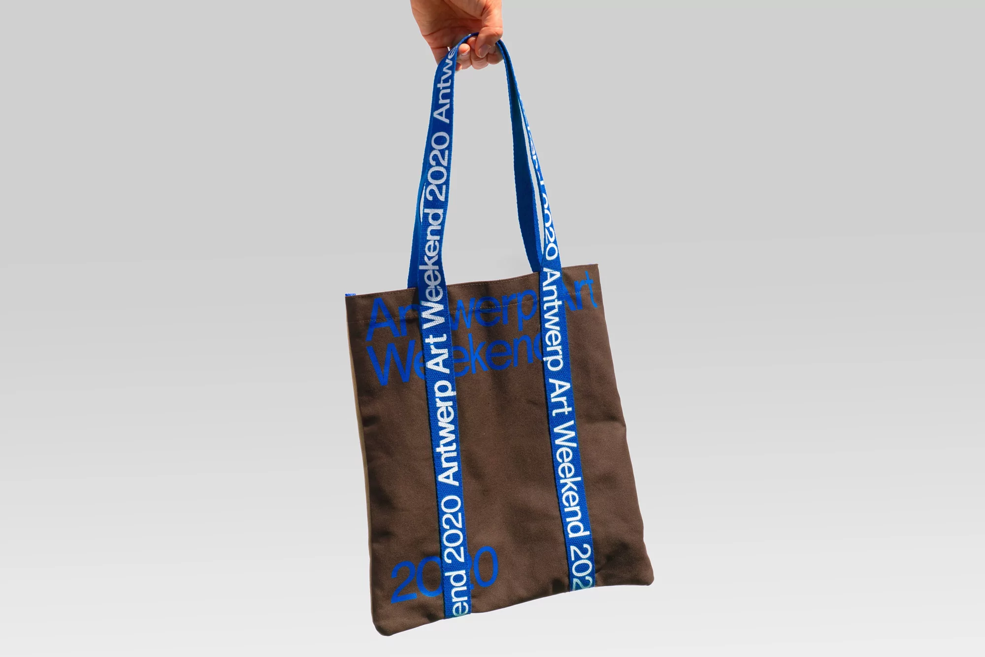 Brown tote bag featuring 'Antwerp Art Weekend 2020' text, suspended by a hand against a light gray backdrop. Ideal for art events.