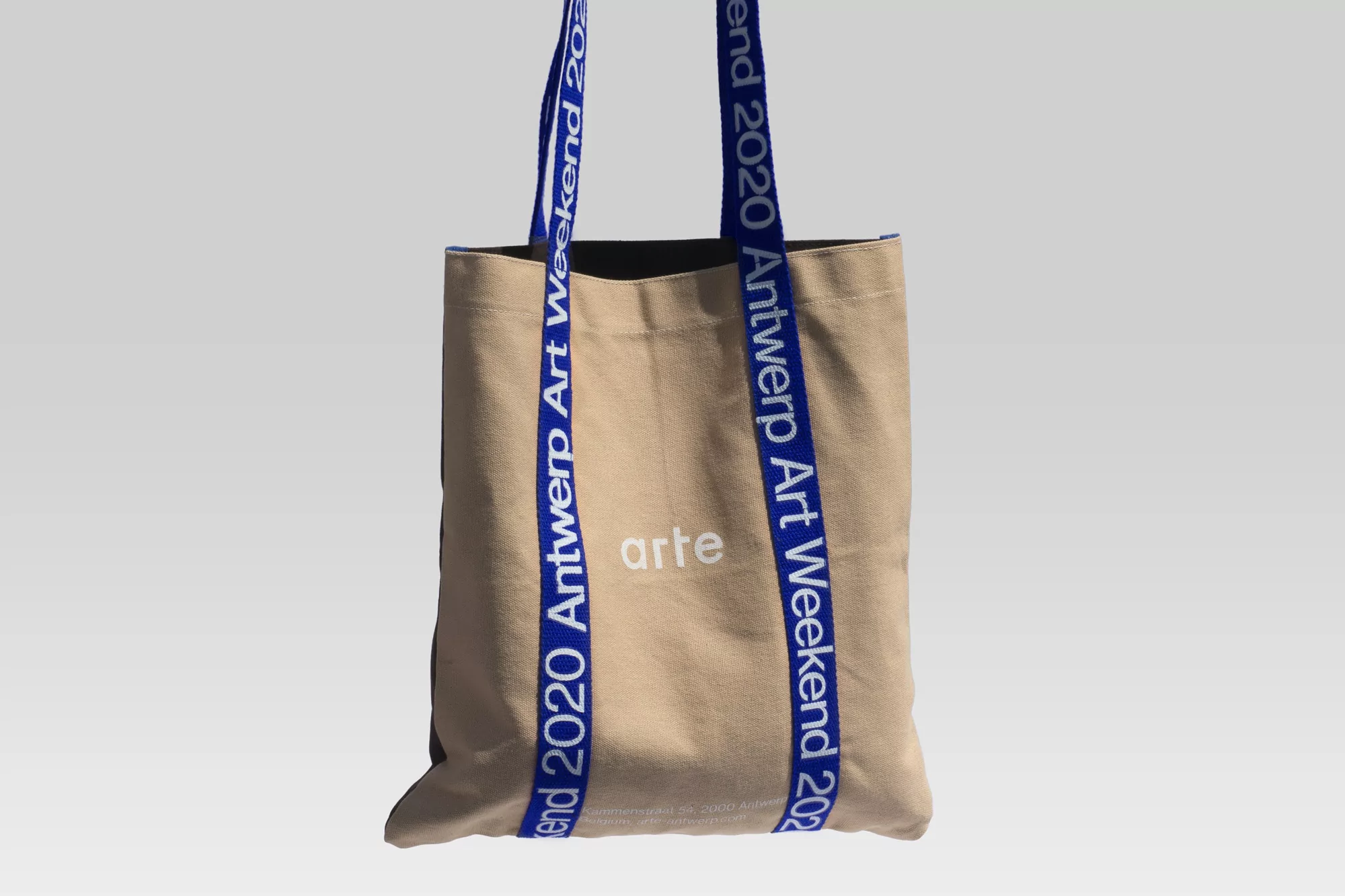 Beige canvas tote bag with blue straps featuring the 'Antwerp Art Weekend 2020' logo, showcasing a simple and stylish design.