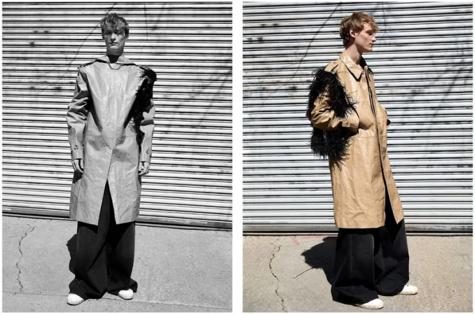 Male fashion model poses in a tan trench coat with black feather accents and wide-leg black pants in front of a metal roll-up door.