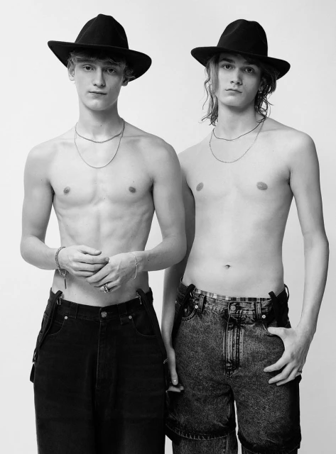 Two shirtless young men in black hats and jeans pose in a studio. One man wears dark jeans with suspenders, the other has light-wash jeans.