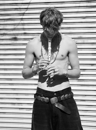 Black and white portrait of a shirtless man with long hair, playing a wind instrument while standing in front of a roller door.