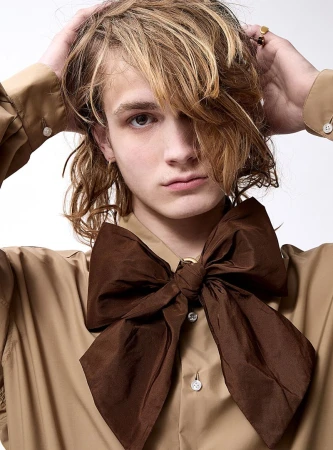 Portrait of a young adult with shoulder-length wavy blonde hair, wearing a tan button-down shirt and an oversized brown satin bow tie.