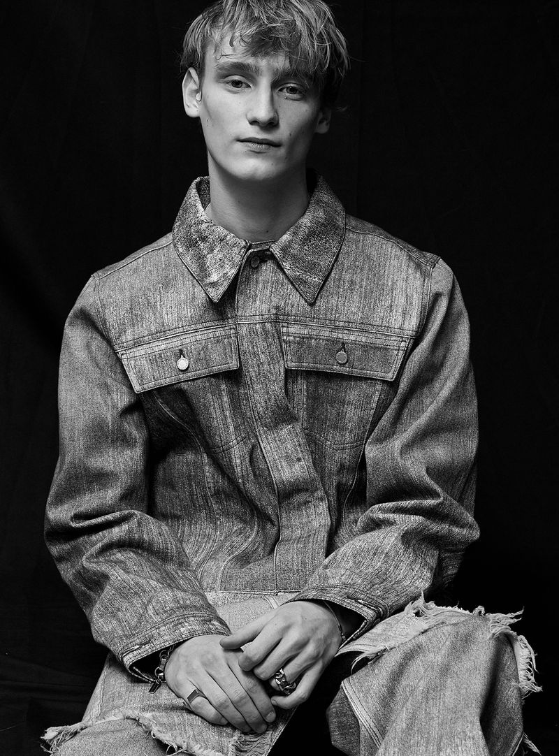 Black and white portrait of a young adult male sitting and wearing a denim jacket, looking directly at the camera with a neutral expression.