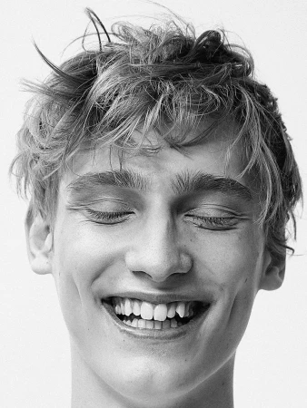 Black-and-white headshot of a young adult man with tousled hair, eyes closed, and a beaming smile, set against a plain white backdrop.