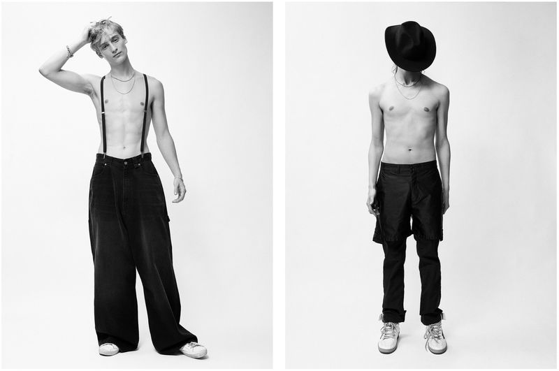Two shirtless adult men in black pants pose in a minimalist studio with a white background, showcasing fashion and style in a monochrome setting.
