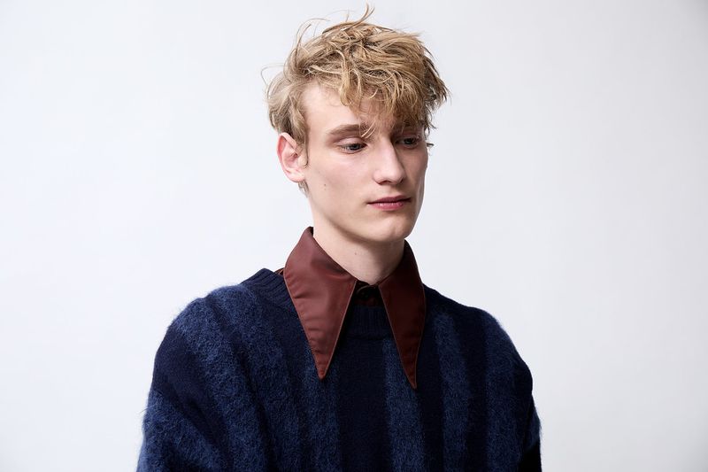 Portrait of a blonde adult male in a striped blue sweater with a brown collar against a white background, looking off-camera with a serious expression.