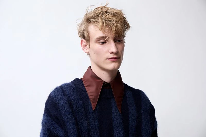 Portrait of a blonde adult male in a striped blue sweater with a brown collar against a white background, looking off-camera with a serious expression.