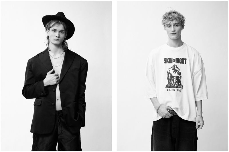 Two young adult men pose indoors against a white backdrop. The left model wears a black hat and blazer, the right wears a graphic t-shirt.