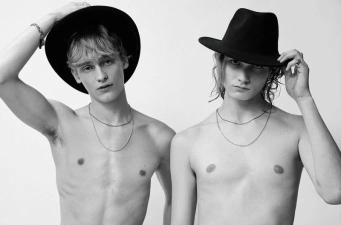 Black and white portrait of two shirtless young adult men wearing hats, posing in front of a plain white background.