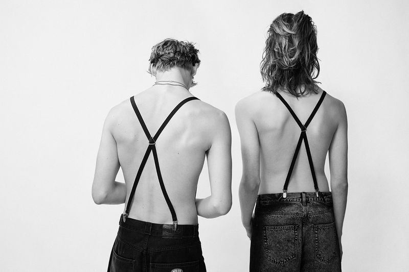 Two people stand against a white background. The people are wearing dark jeans and suspenders. The suspenders are black and thin.