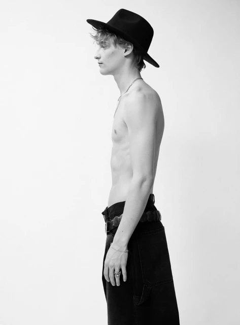 Black and white studio shot of a shirtless young adult man wearing a fedora, black jeans, and a decorative belt against a white backdrop.
