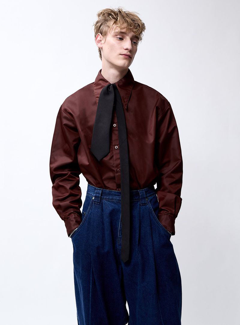 Studio shot of a young adult male model wearing a shiny dark red button-down shirt with a black tie and baggy dark blue denim pants.