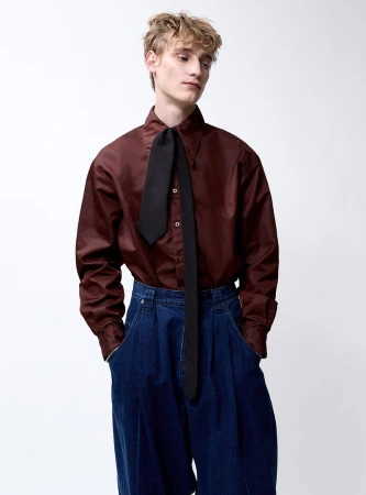 Studio shot of a young adult male model wearing a shiny dark red button-down shirt with a black tie and baggy dark blue denim pants.