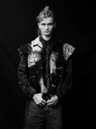 Black and white portrait of a young adult man in a denim vest over a plaid shirt, shot in a photo studio with a dark background.