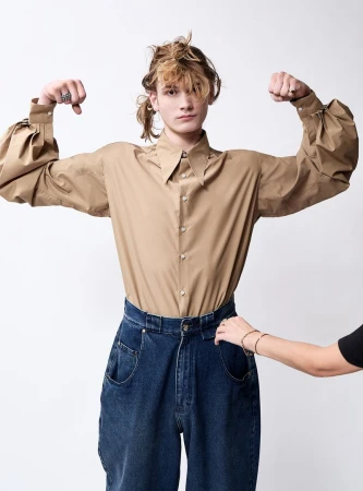 Young adult man in a tan shirt and wide-legged jeans with his arms flexed in a studio, another person adjusting the jeans' waistband.