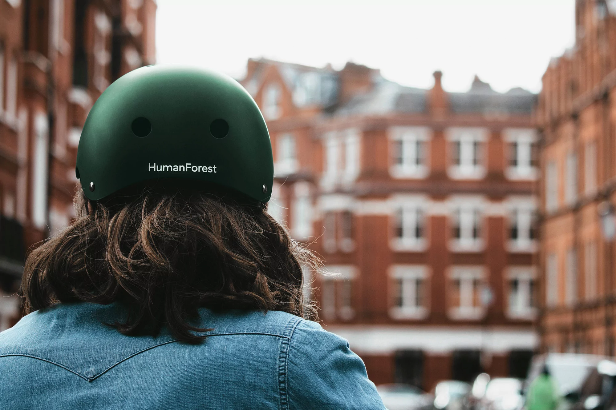 Person wearing a green HumanForest bicycle helmet and a denim jacket walking in an urban environment with blurred buildings.